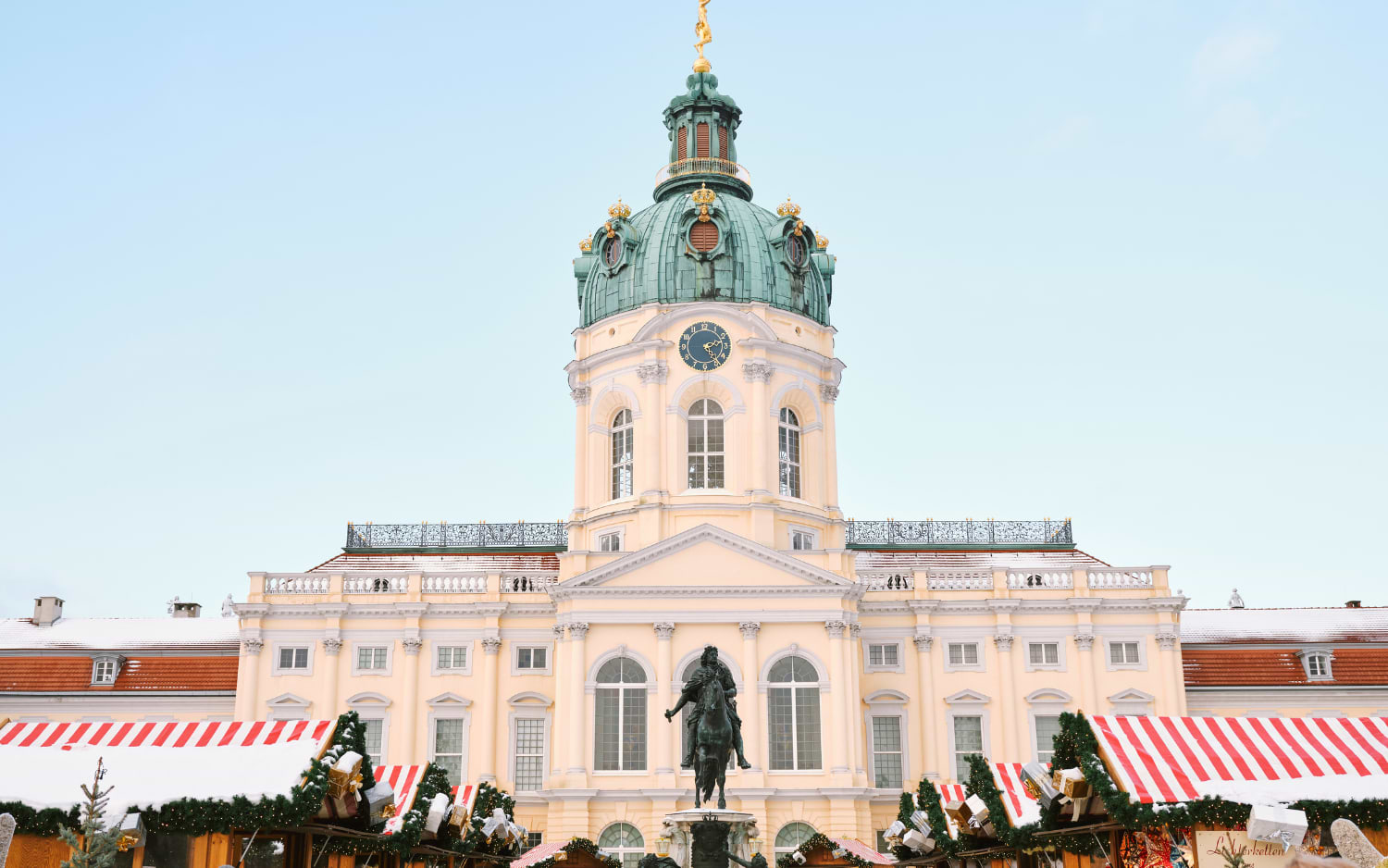 Christmas market stalls with red-and-white roofs in front of snowy Charlottenburg Palace and its green dome in Berlin