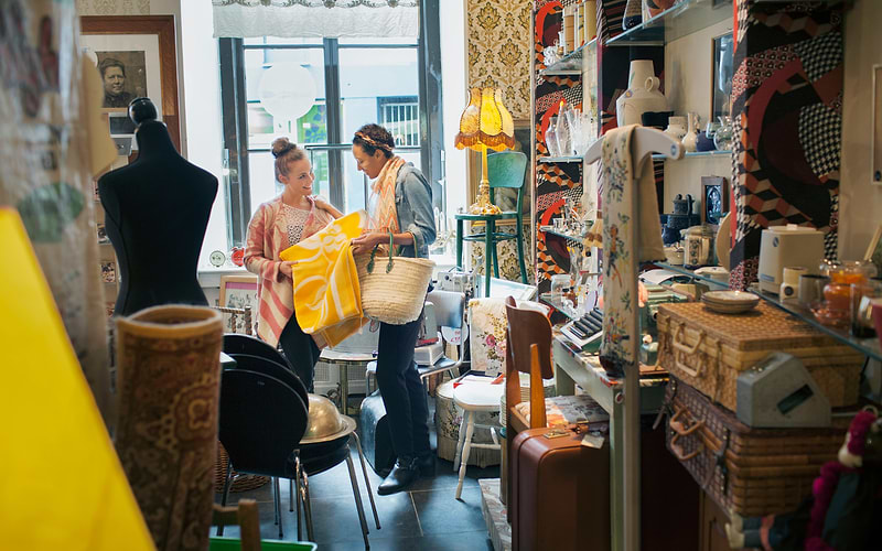 Two women examine a yellow blanket in a cluttered antique shop full of lamps, cases and ceramics