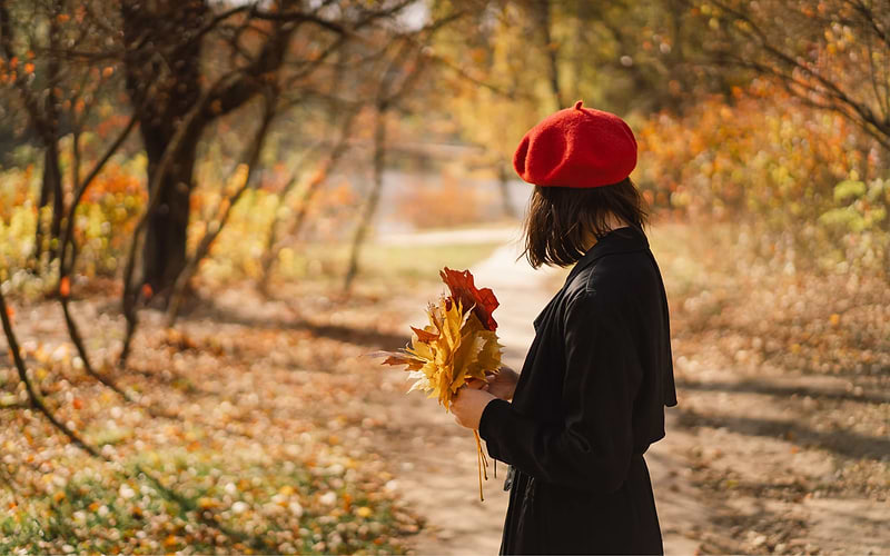 Woman in a red beret and black coat holding a bouquet of golden maple leaves on a park path, a classic autumn travel scene