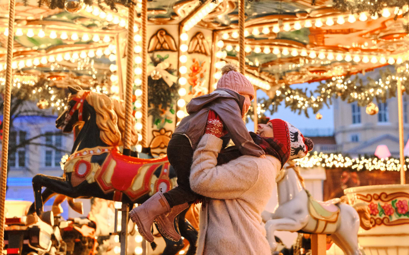 Mother lifts her child onto a lit carousel with painted horses at a Berlin Christmas market in winter