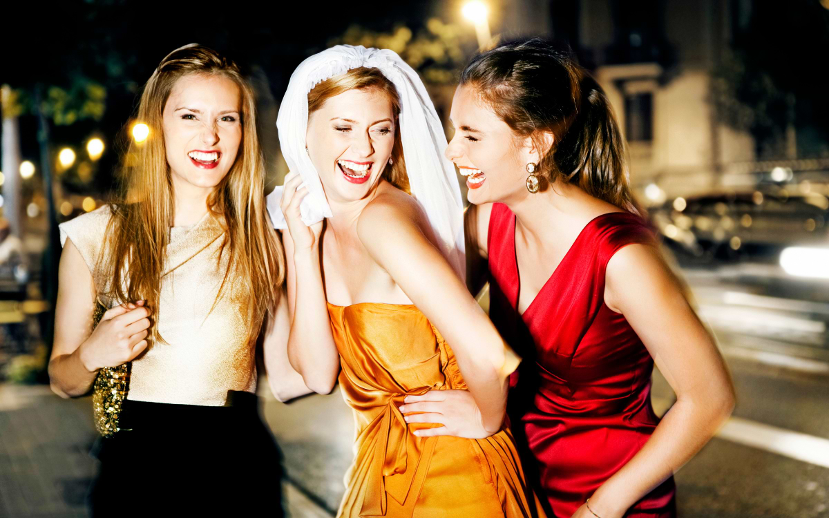 Bride wearing a veil laughing with two friends in evening dresses on a lit-up city street at night, hen party Europe