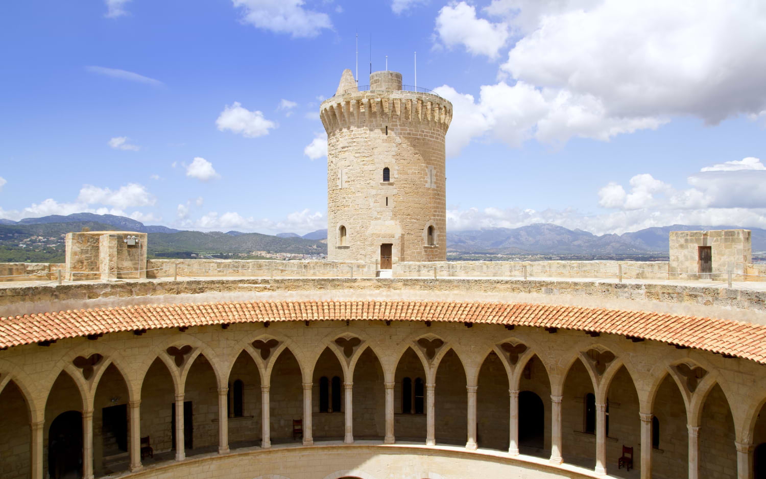 The round tower of Bellver Castle overlooks its Gothic cloister courtyard in Palma, with the Tramuntana mountains visible in the background.