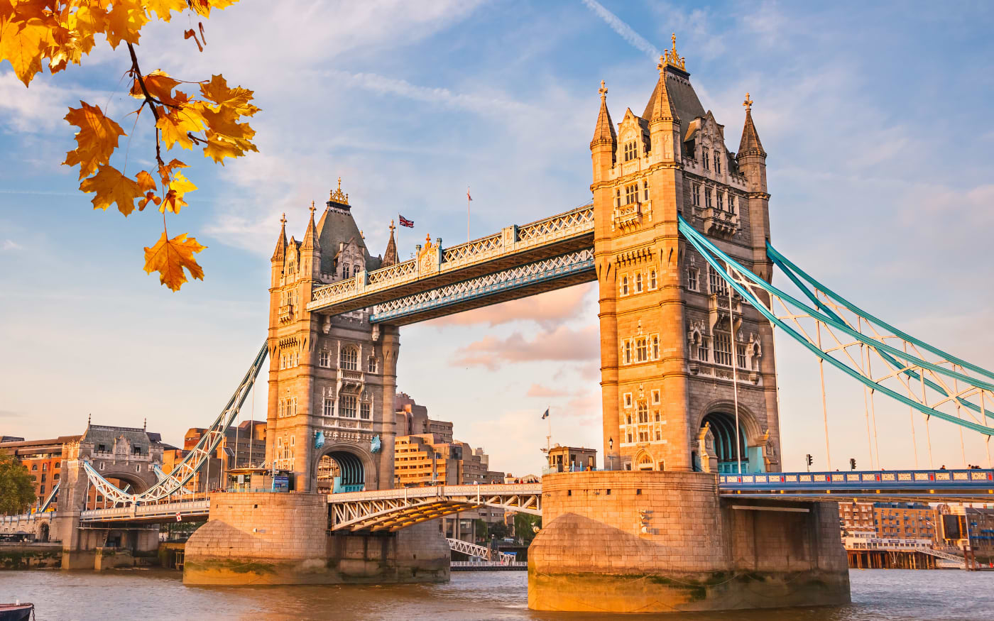 Tower Bridge in London glowing in golden evening light over the Thames, framed by yellow autumn maple leaves