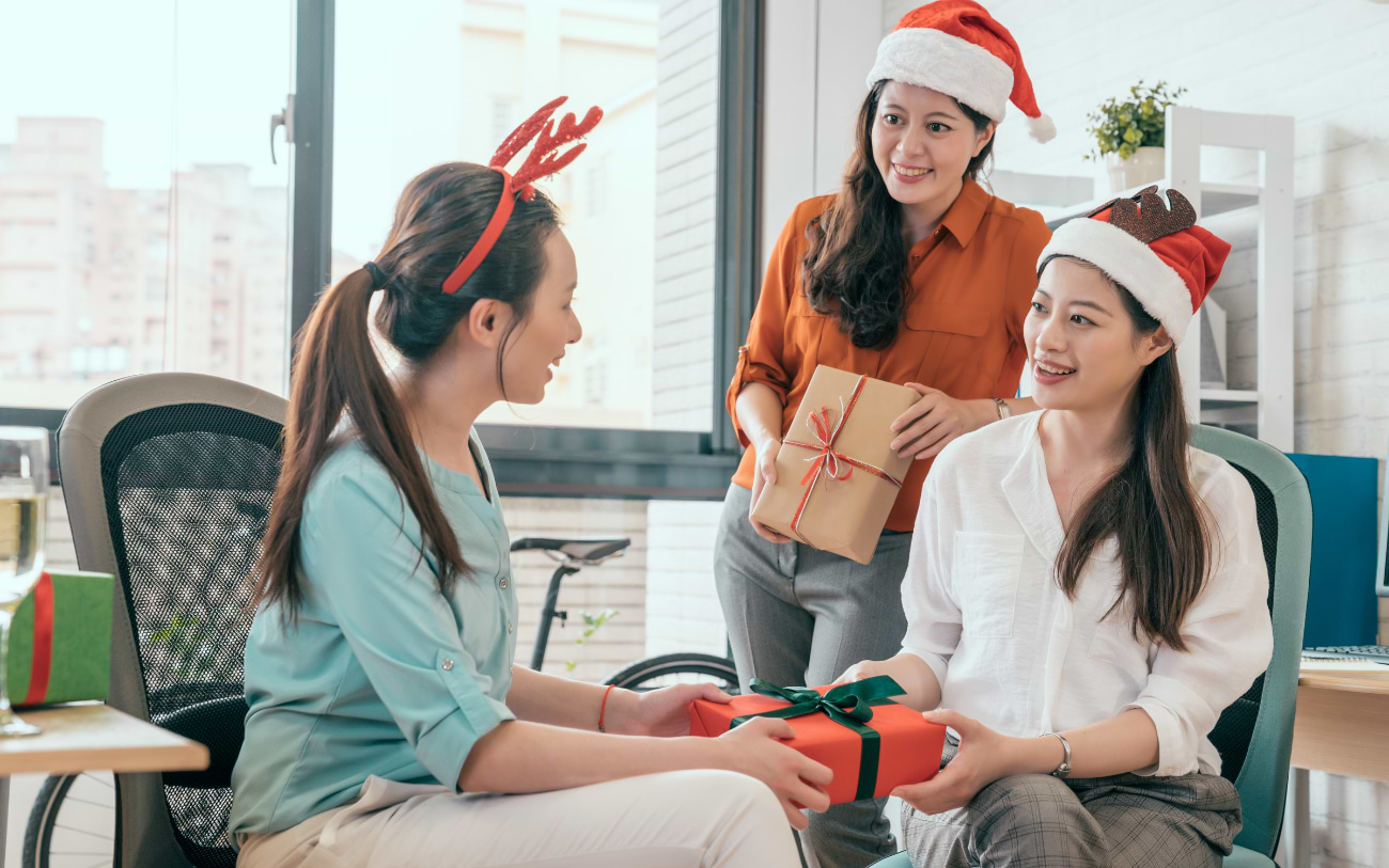 Drei Kolleginnen mit Weihnachtsmützen und Rentiergeweih tauschen im Büro verpackte Geschenke aus, ein fröhlicher Wichtelmoment am Fenster