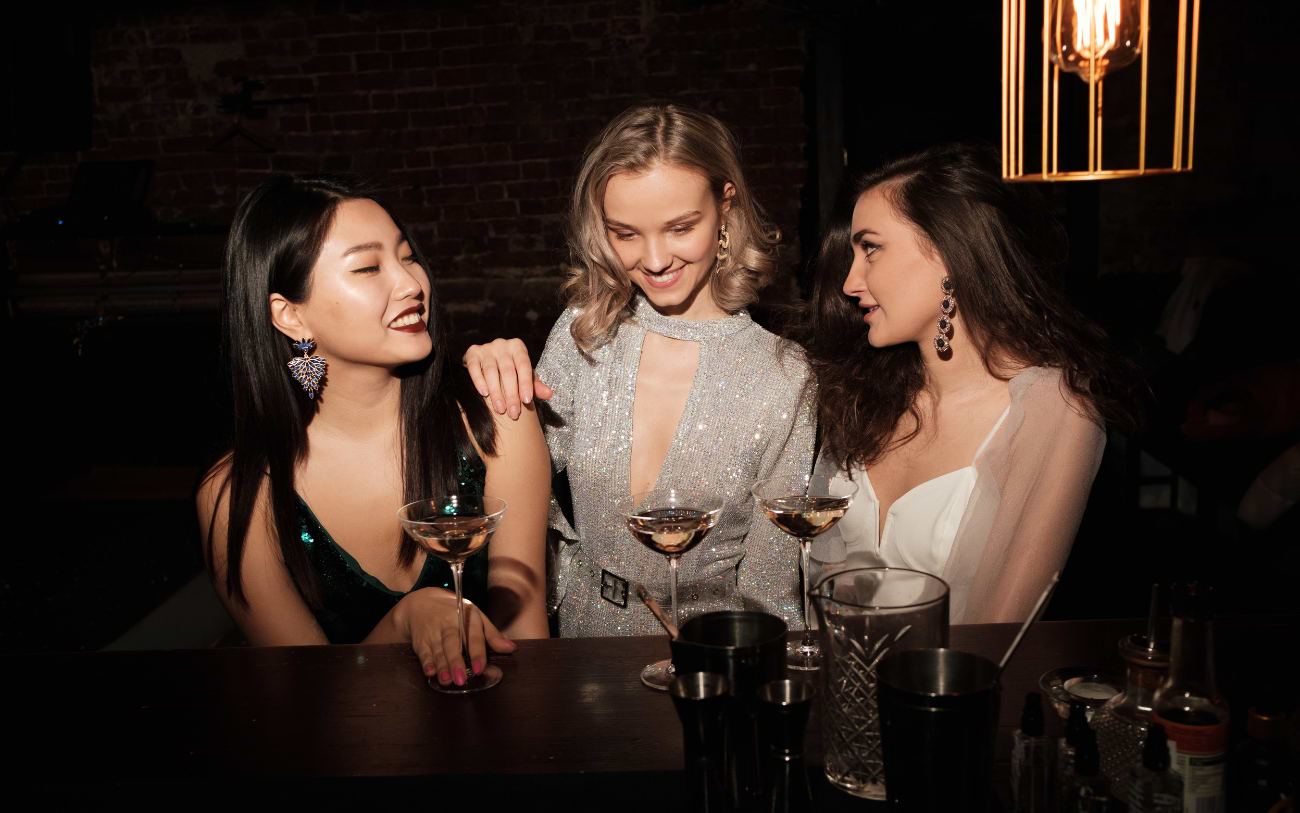Three elegantly dressed women smiling with coupe glasses at a dark wooden bar counter under warm lighting