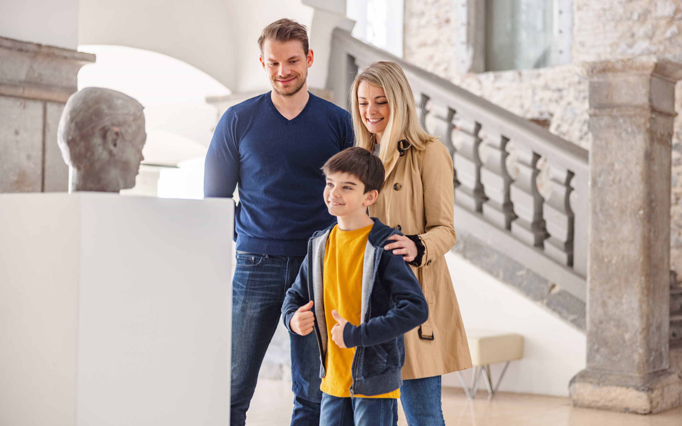 A father, mother and young son smiling as they look at a bronze head sculpture during a family museum visit in Munich, Germany.
