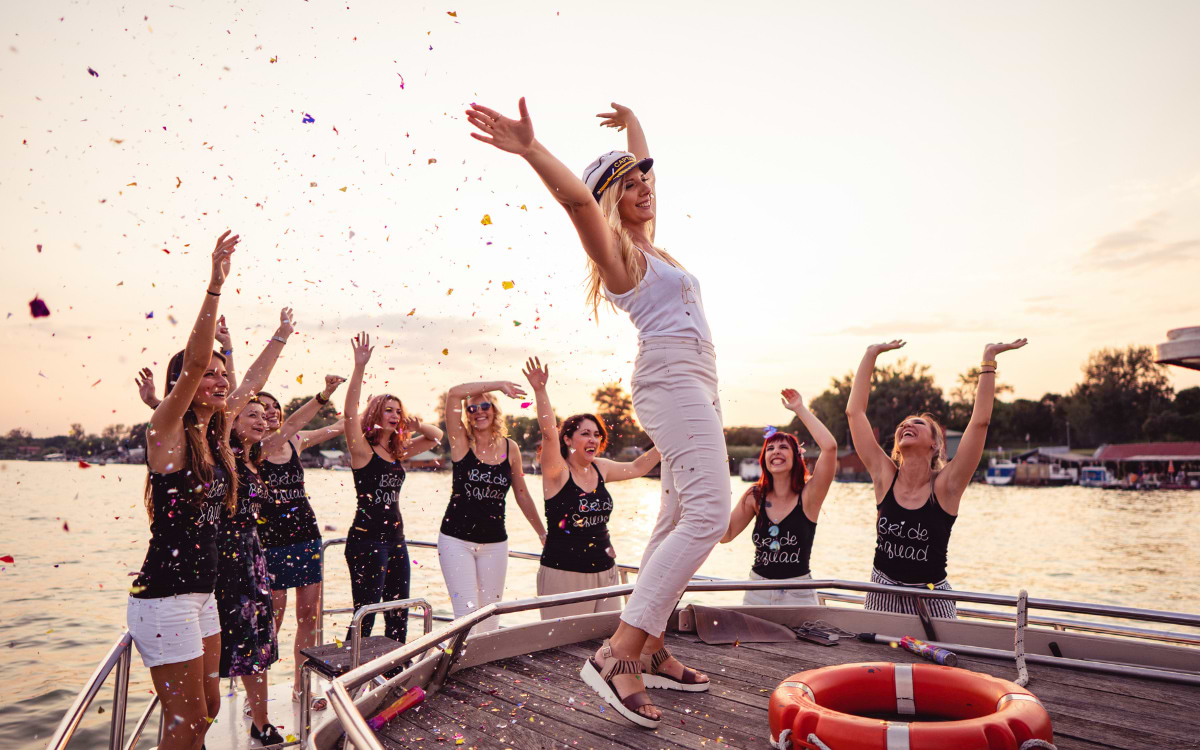 Bride in white outfit and captain's hat celebrating with her hen party squad on a boat at sunset, confetti flying