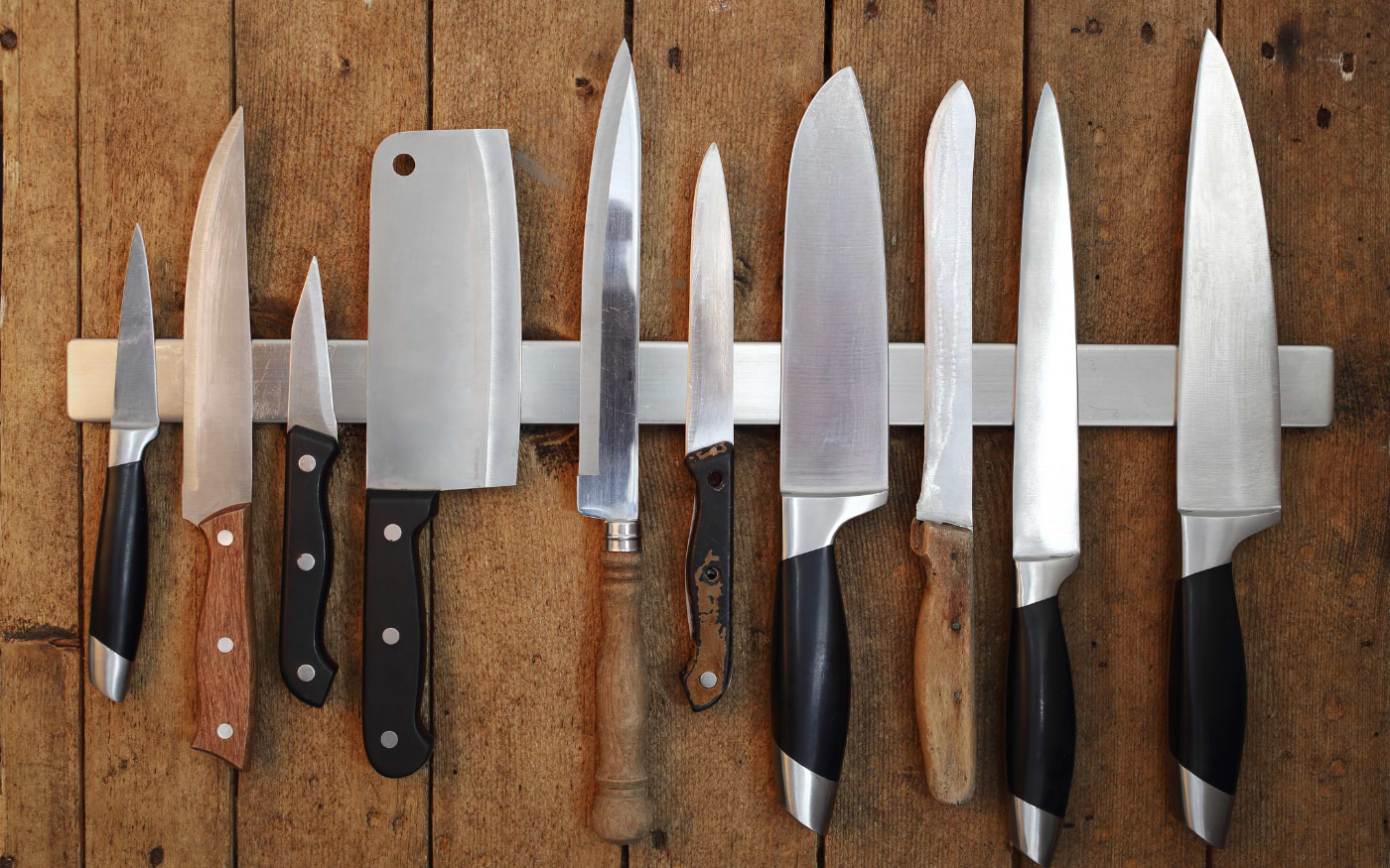 Set of kitchen knives of varying sizes hanging on a magnetic strip against a wooden wall
