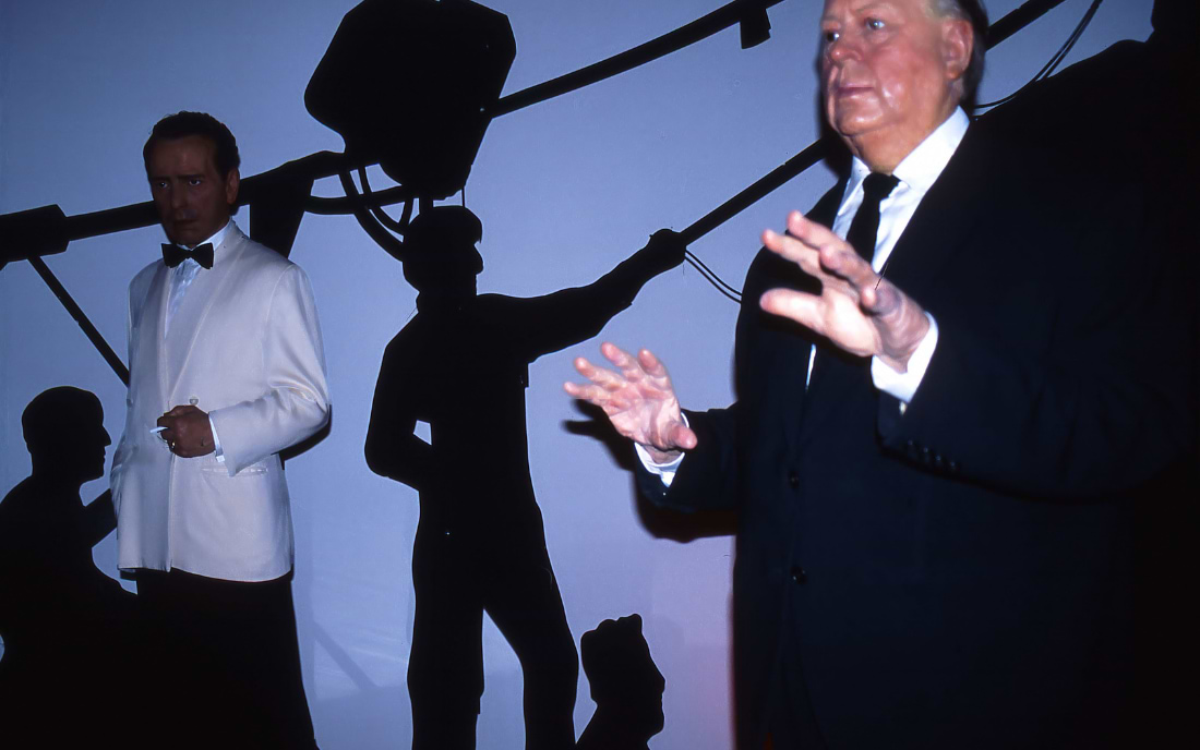 Lifelike wax figures of a man in a white tuxedo and an older gentleman in a dark suit, displayed against a dramatic blue-lit film set backdrop with studio lighting equipment at Madame Tussauds in London