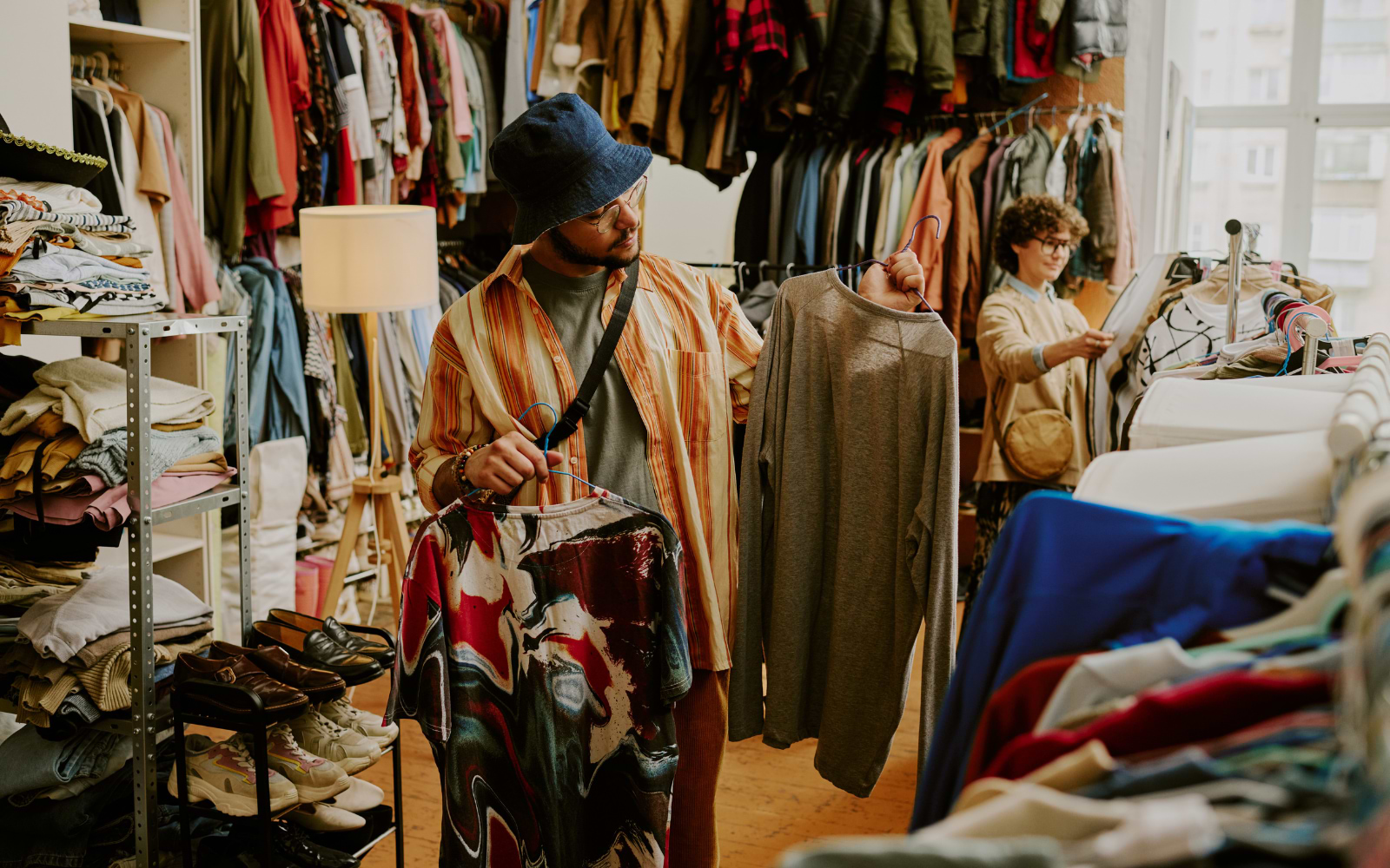 Man in a bucket hat compares two shirts on hangers while browsing racks in a busy second hand shop