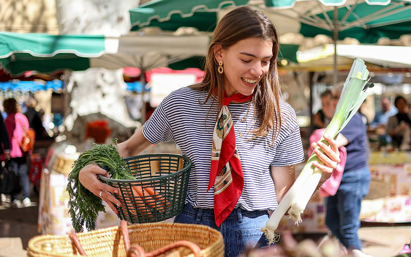 A smiling shopper holds fresh carrots and a leek at a lively Barcelona outdoor market stand.