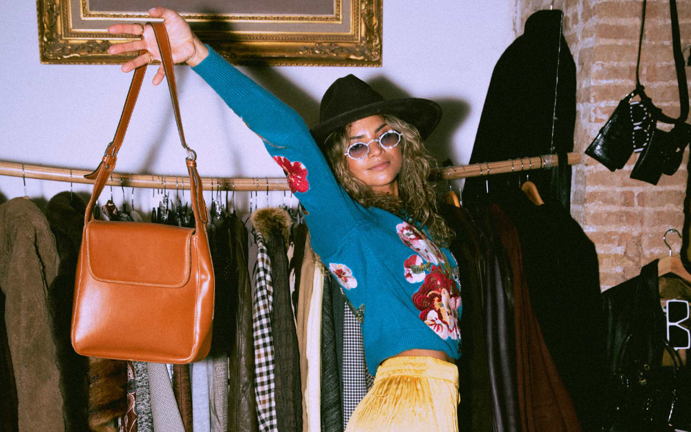 Woman holding up a leather vintage handbag while browsing clothing racks at a vintage market