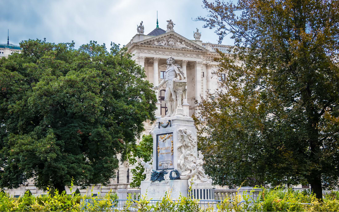 White marble Mozart Monument in Vienna's Burggarten, framed by trees, with the Hofburg facade in the background
