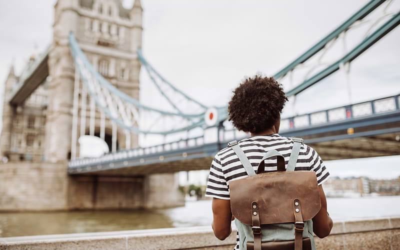 Traveller with a leather backpack and striped T-shirt looking at Tower Bridge from the Thames riverside in London